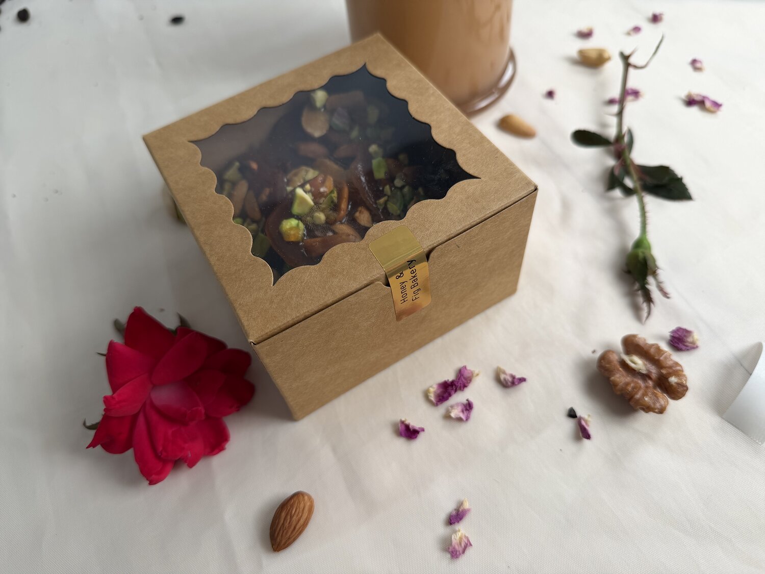 A handmade kraft gift box of honey mixed-nut brittle, sealed with a Honey & Fig Bakery gold sticker, with a rose and nuts on linen — Pflugerville, Texas