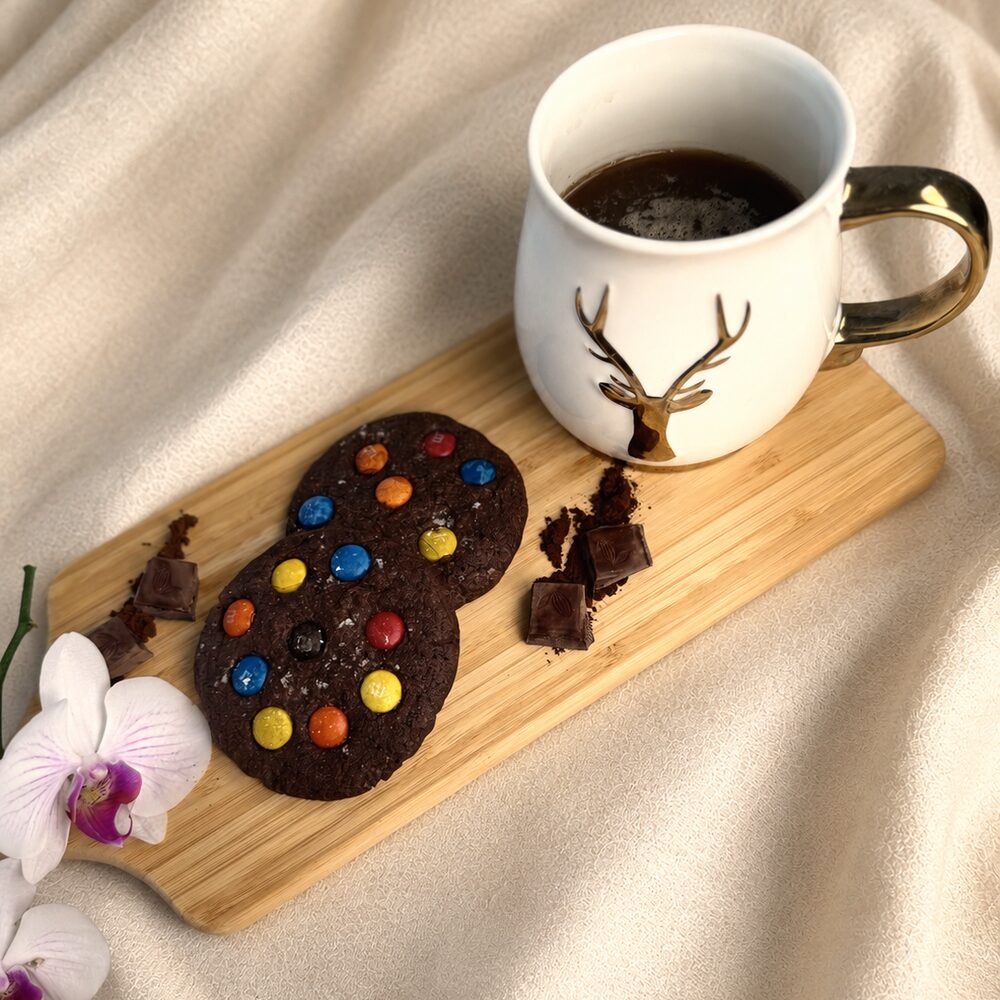 Two dark chocolate cookies studded with colorful candy pieces on a wooden board beside a cup of coffee, Honey & Fig Bakery, Pflugerville, Texas
