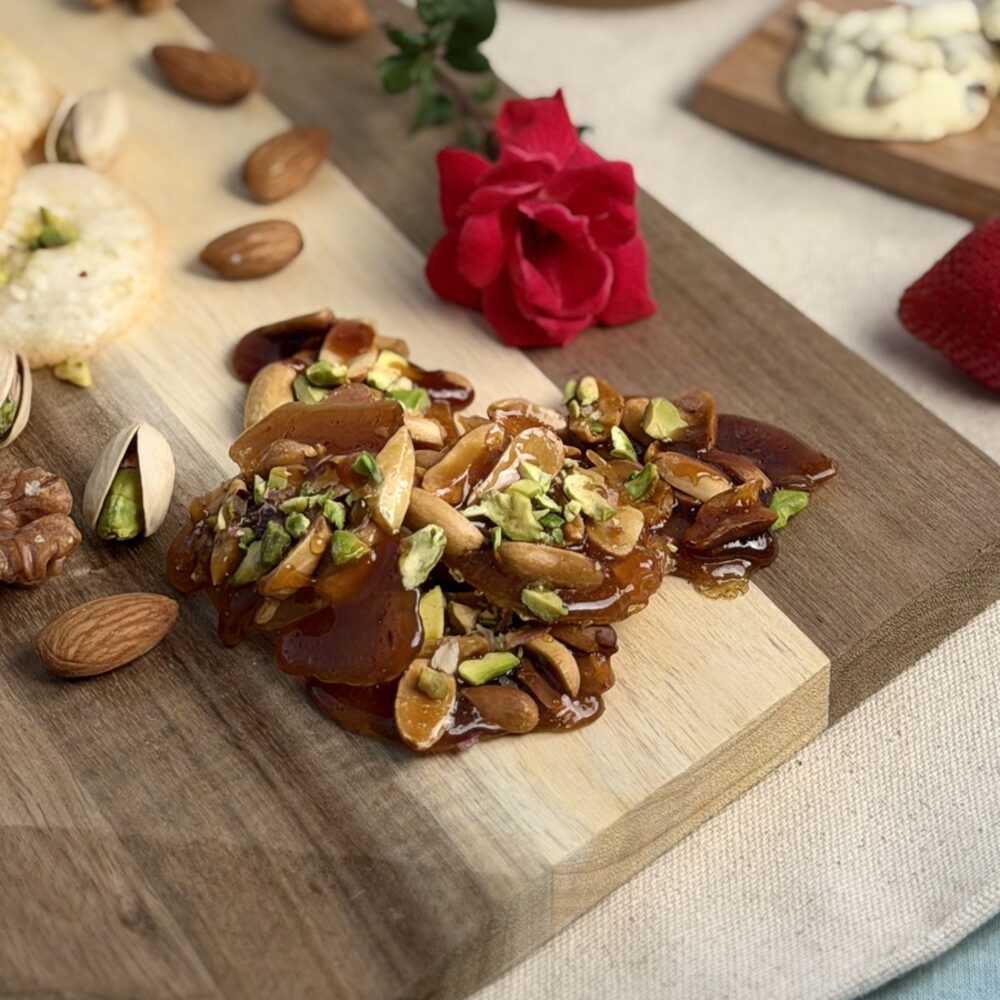 Glossy almond and pistachio brittle on a wooden board with whole almonds and a red rose, Honey & Fig Bakery, Pflugerville, Texas